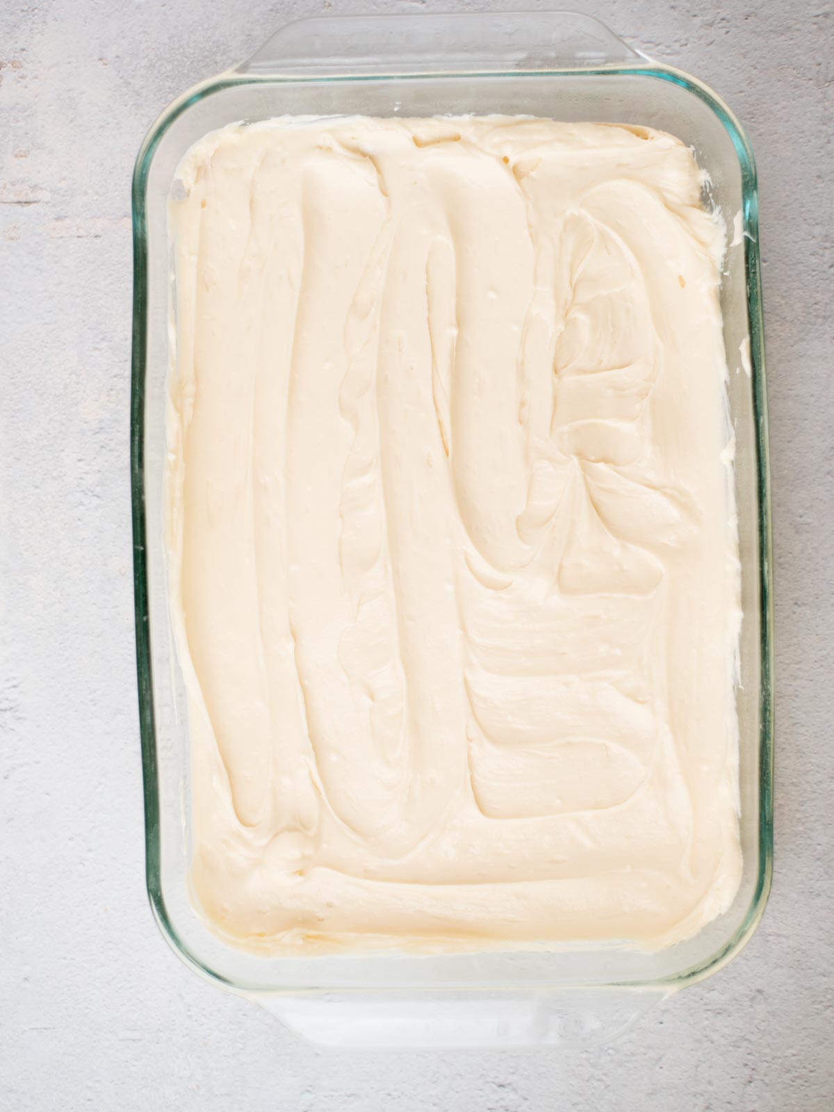A glass baking dish filled with smooth, creamy white frosting spread evenly across its surface. The dish is set on a light gray textured background.
