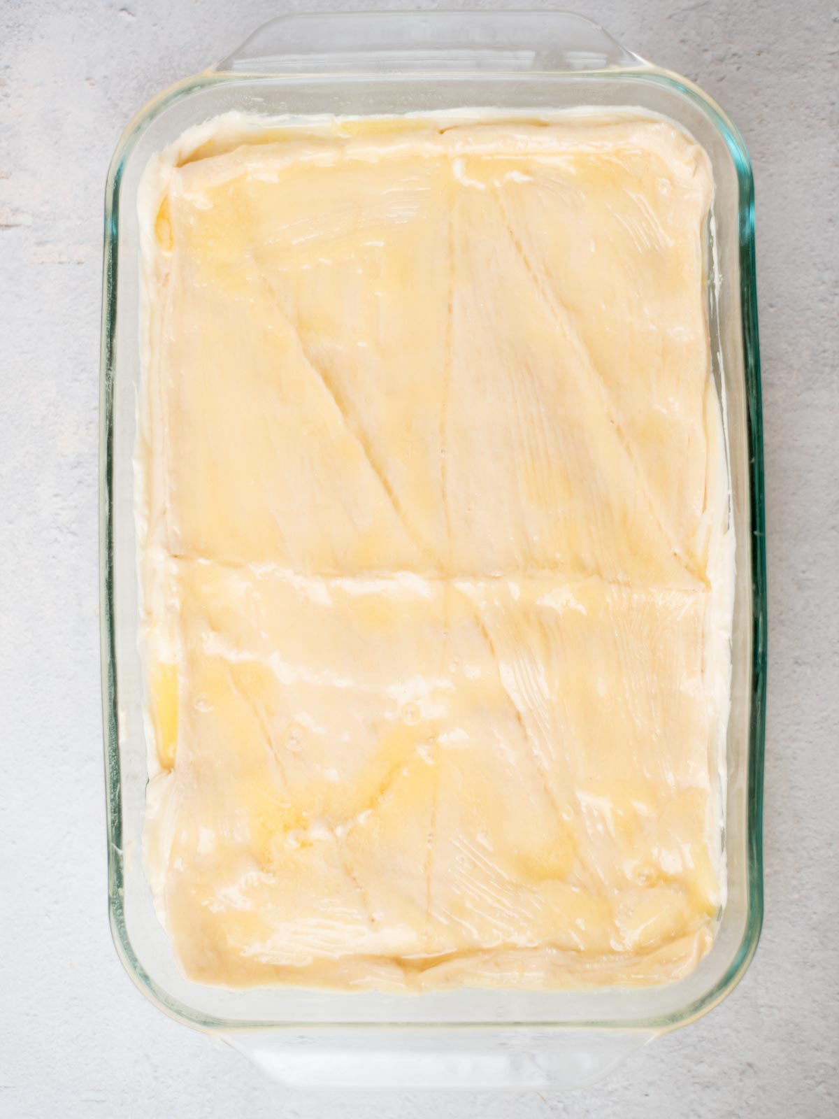 A glass baking dish filled with unbaked dough, scored with diagonal lines. The dough is golden and smooth, ready for baking. The background is a light gray surface.