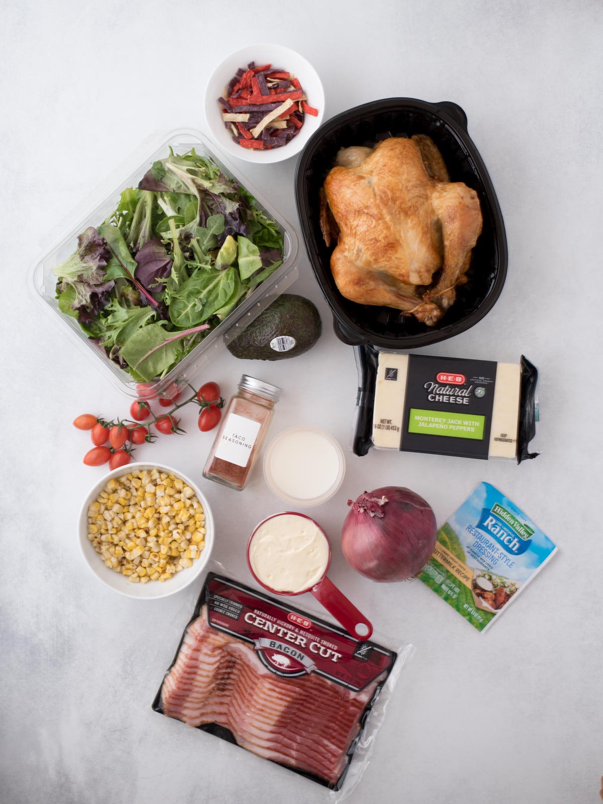 A flat lay of assorted food items including a container of mixed greens, a cooked whole chicken, cherry tomatoes, an avocado, block of cheese, salad dressing, red onion, spices, corn, Ranch seasoning, and a package of bacon on a light surface.