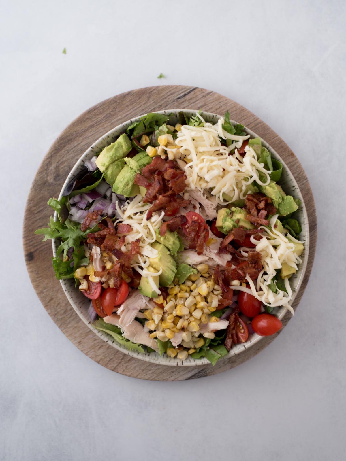 A bowl of salad on a wooden plate, containing shredded cheese, diced avocado, corn, bacon bits, cherry tomatoes, lettuce, and red onion slices.