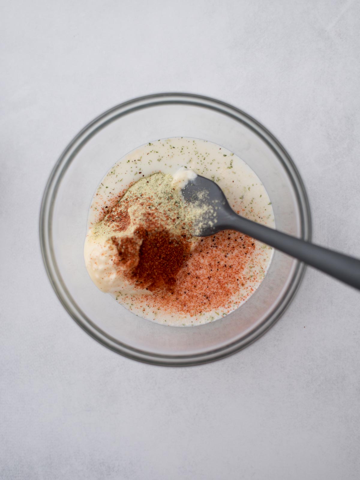 A mixing bowl with a creamy white sauce being combined with various spices, including red and green powders. A black spoon is in the bowl, suggesting the ingredients are being blended. The background is a light gray surface.