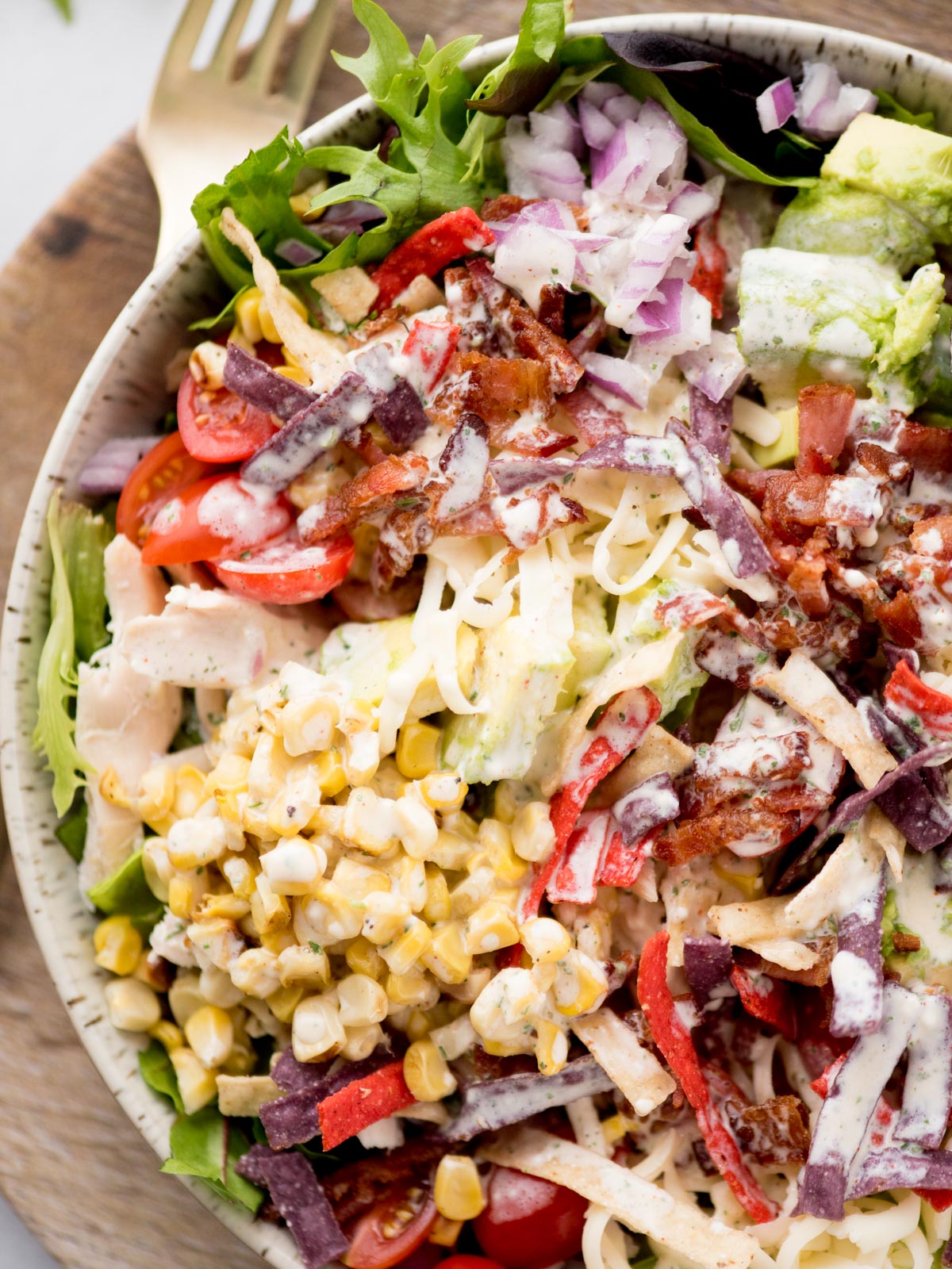A colorful salad in a bowl with mixed greens, cherry tomatoes, grilled corn, shredded cheese, avocado, red onions, tortilla strips, and bacon, topped with creamy dressing. A fork is placed beside the bowl on a wooden surface.