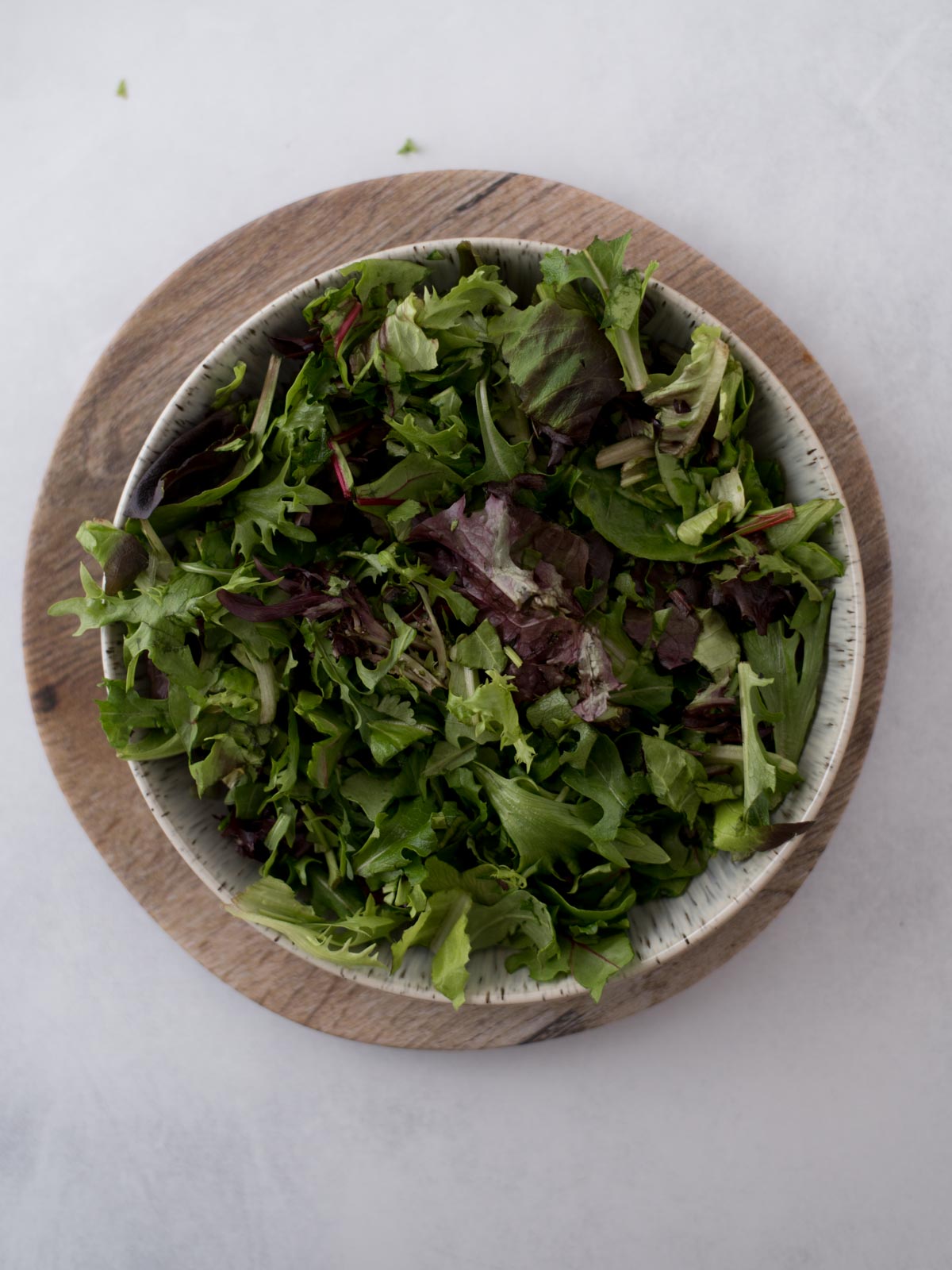 A bowl of mixed leafy greens sits on a wooden board. The greens vary in shades of green and purple, with different textures and shapes, creating a fresh and vibrant appearance against a light background.