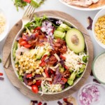 A vibrant salad in a bowl on a wooden plate, featuring diced avocado, shredded cheese, cherry tomatoes, corn, bacon, tortilla strips, and sliced red onions, garnished with a halved avocado. A fork is placed beside the bowl.