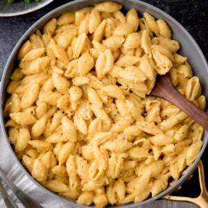 A skillet filled with creamy mac and cheese pasta shells, garnished with cracked black pepper. A wooden spoon rests in the dish, ready to serve. The background shows a hint of a green leafy side dish.