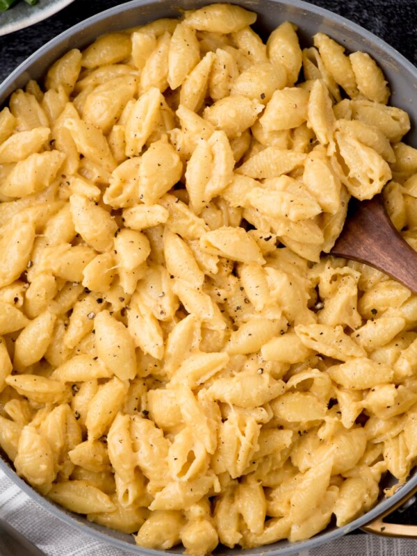 A skillet filled with creamy mac and cheese pasta shells, garnished with cracked black pepper. A wooden spoon rests in the dish, ready to serve. The background shows a hint of a green leafy side dish.