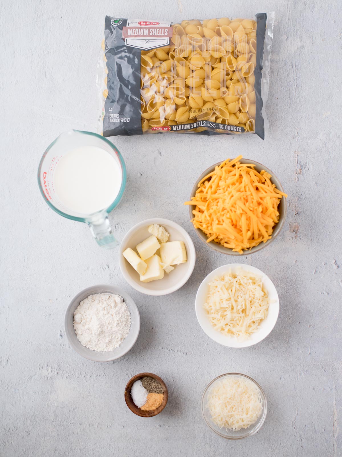 Top view of ingredients for macaroni and cheese on a light surface. Visible items include a bag of medium shell pasta, shredded cheddar cheese, grated Parmesan cheese, butter, flour, a measuring cup of milk, and small bowls of salt and nutmeg.