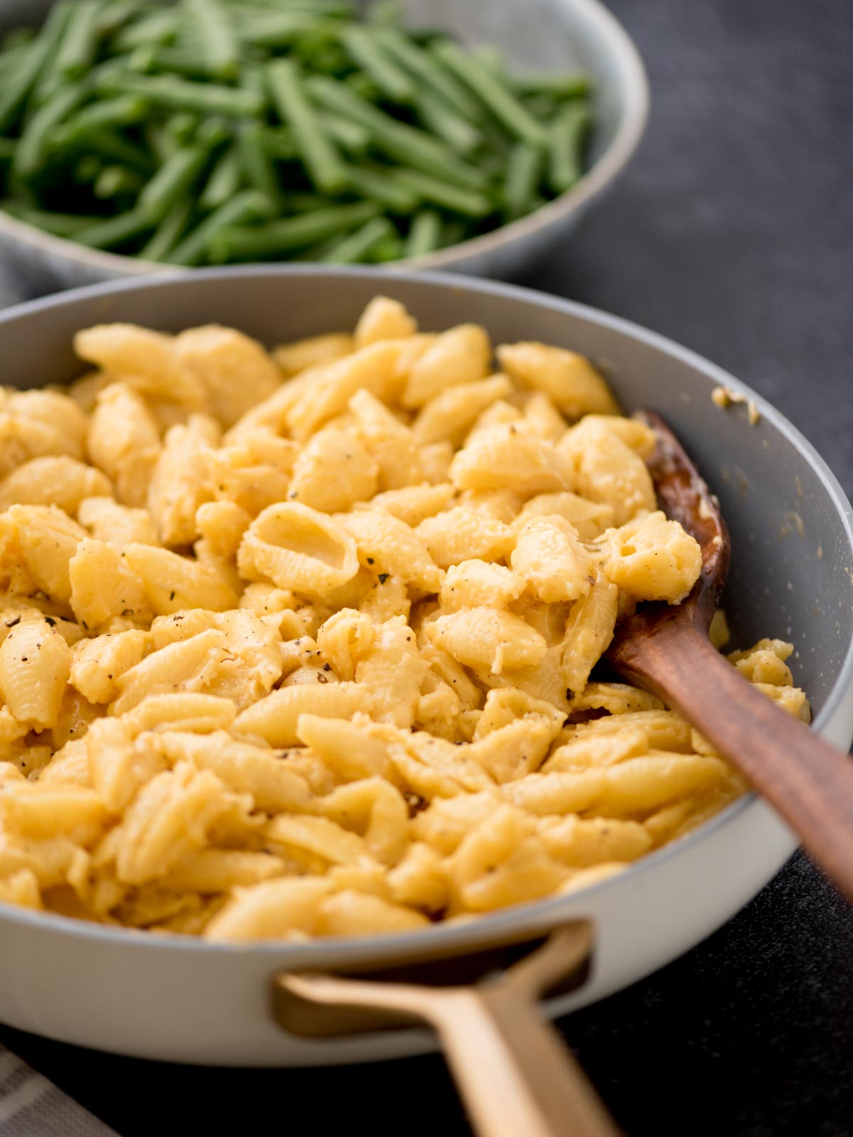 A skillet filled with creamy macaroni and cheese, with a wooden spoon resting inside. In the background, a bowl of fresh green beans is partially visible. Both dishes are placed on a dark surface.