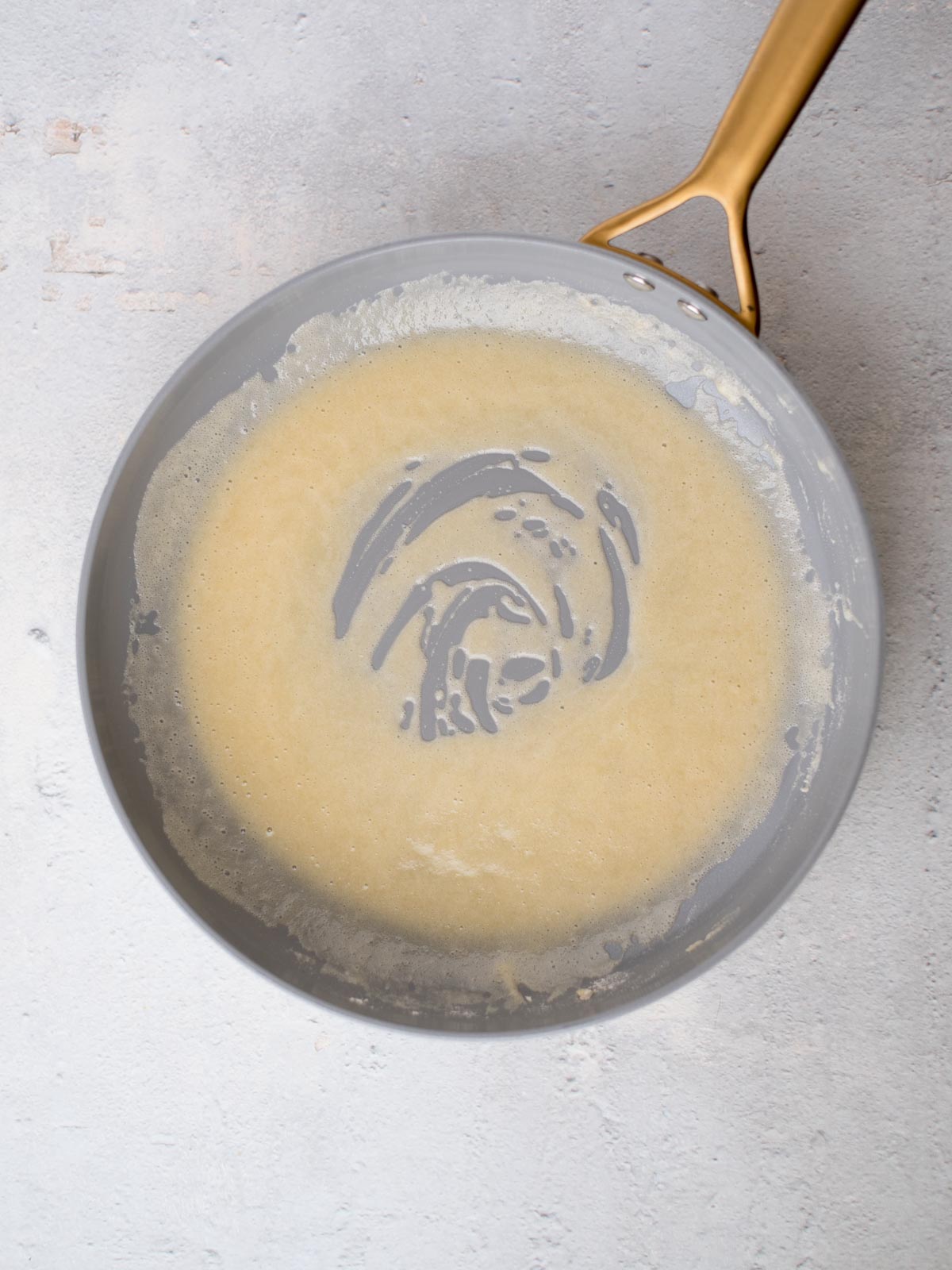 A non-stick frying pan with a swirl of beige batter or sauce on a light gray, textured surface. The handle is metallic and golden-colored.