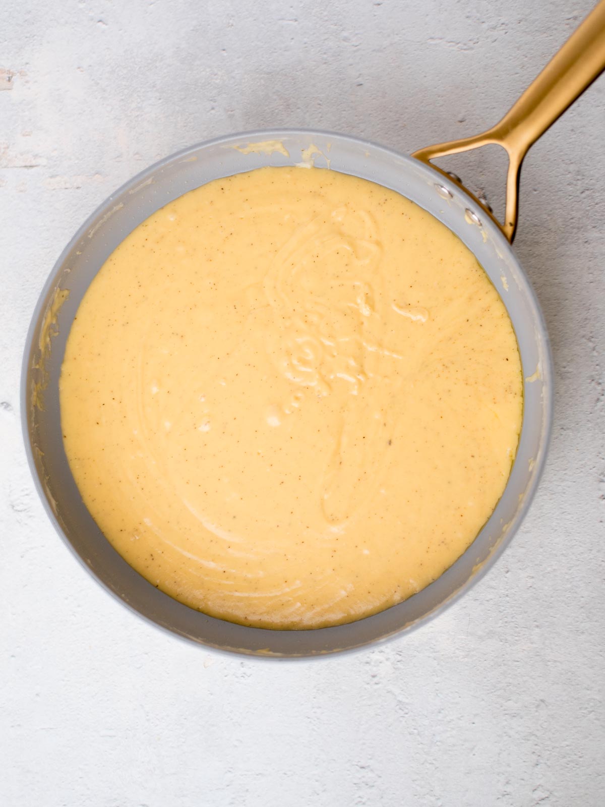 A round pan filled with a creamy, yellowish batter sits on a light gray surface. The batter appears smooth and slightly thick, with visible specks, possibly indicating spices or seasoning. The pan has a brass-colored handle.