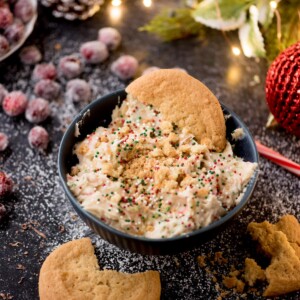 A dark bowl filled with cookie dough topped with colorful sprinkles, surrounded by crumbled cookies. Frosted cranberries and holiday decorations, including a pine cone and red ornament, are in the background.