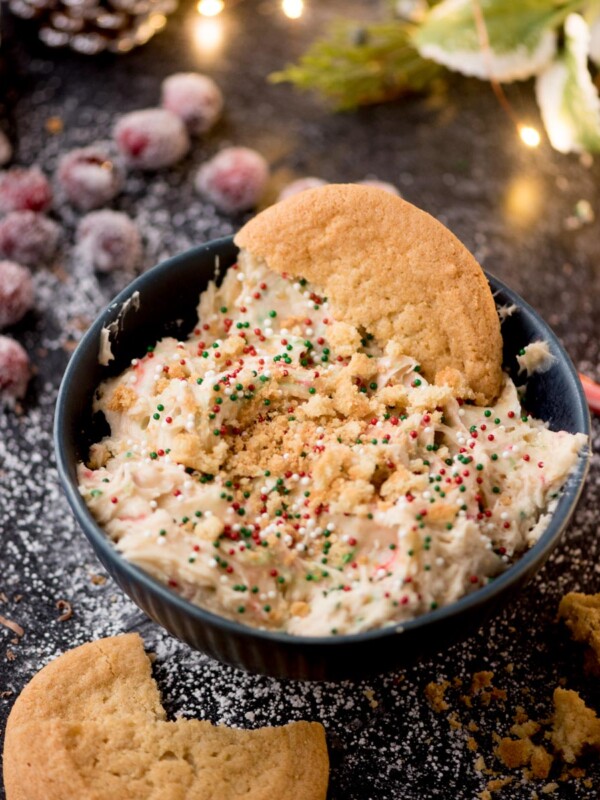 A dark bowl filled with cookie dough topped with colorful sprinkles, surrounded by crumbled cookies. Frosted cranberries and holiday decorations, including a pine cone and red ornament, are in the background.