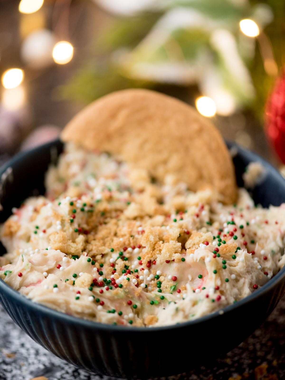 A bowl of creamy, festive dessert dip topped with red, green, and white sprinkles. A cookie is partially dipped into the mixture. Soft lights and blurred holiday decorations are in the background.