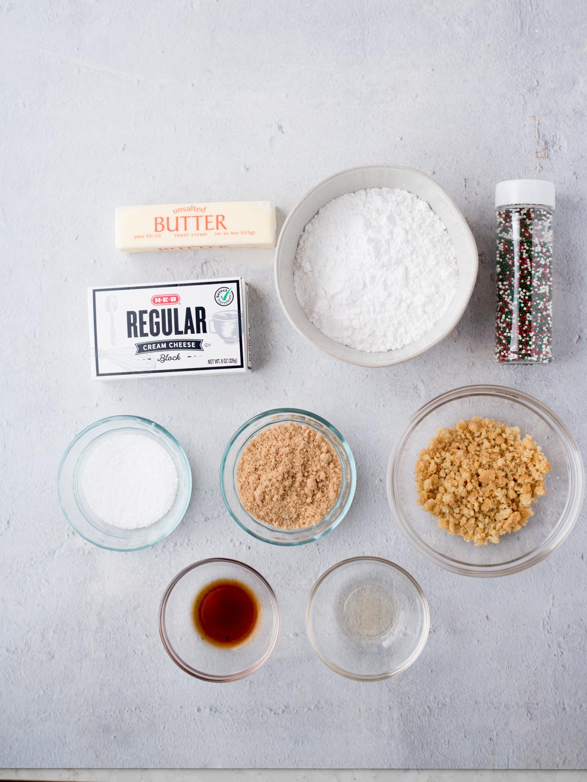 Top-down view of ingredients on a gray surface, including a stick of butter, a box of cream cheese, powdered sugar in a bowl, small bowls of white and brown sugar, vanilla extract, and sprinkles in a clear bottle.