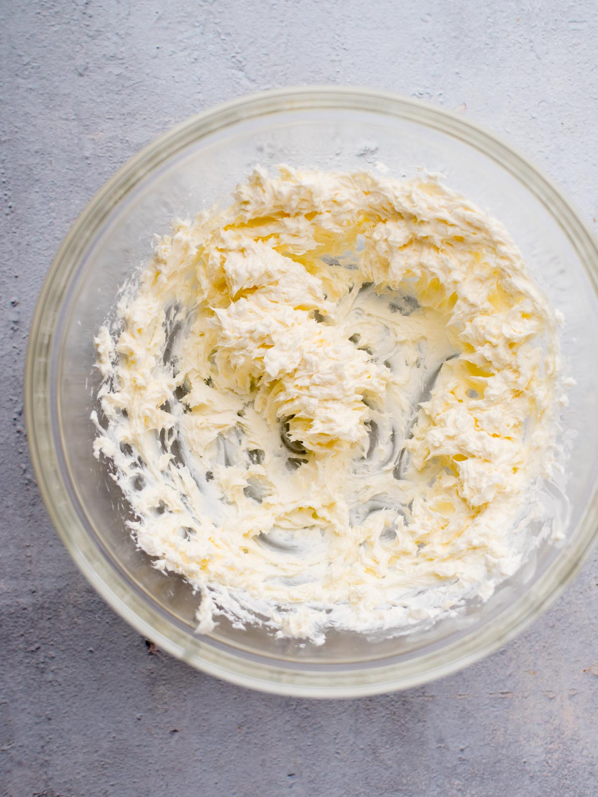 A clear glass bowl containing whipped cream cheese on a gray surface. The cream cheese appears fluffy and smooth, with swirls indicating it has been mixed thoroughly.