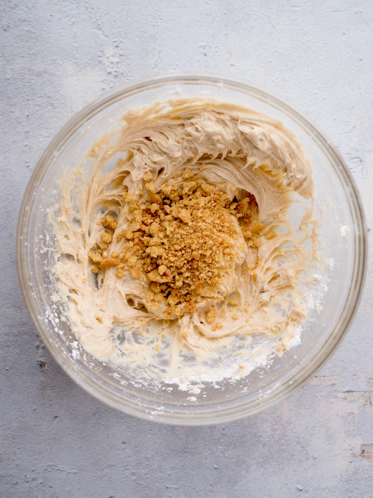 A glass bowl contains a creamy mixture with a heap of crushed nuts or cookie crumbs in the center. The bowl is set on a light gray surface.