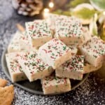 A pile of white fudge squares topped with red, green, and white sprinkles on a dark plate. The background has powdered sugar sprinkled around, with holiday decor and warm lights adding a festive touch.