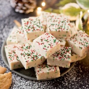 A pile of white fudge squares topped with red, green, and white sprinkles on a dark plate. The background has powdered sugar sprinkled around, with holiday decor and warm lights adding a festive touch.