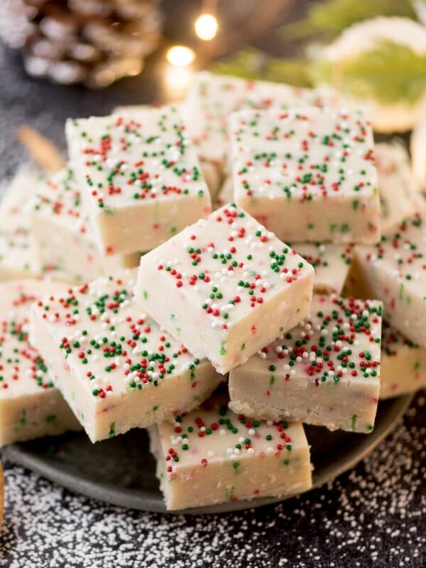 A pile of white fudge squares topped with red, green, and white sprinkles on a dark plate. The background has powdered sugar sprinkled around, with holiday decor and warm lights adding a festive touch.