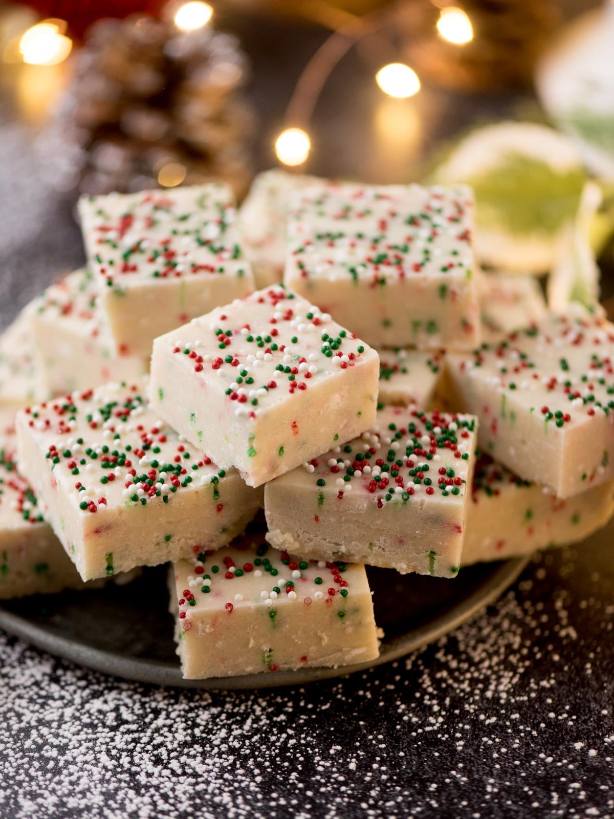 A plate stacked with creamy white fudge squares, topped with festive red and green sprinkles. The background features softly focused holiday lights and a hint of snowy decor.