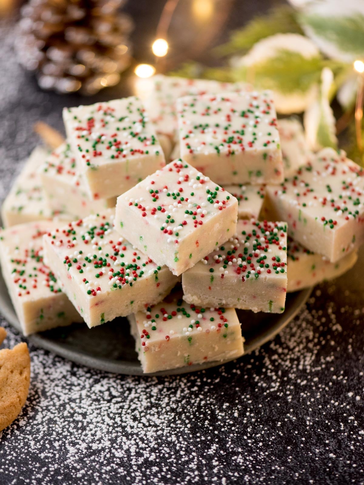 A stack of festive fudge squares on a dark plate, topped with red, green, and white sprinkles. The background features soft holiday lights, a pine cone, and a dusting of powdered sugar, enhancing the seasonal atmosphere.