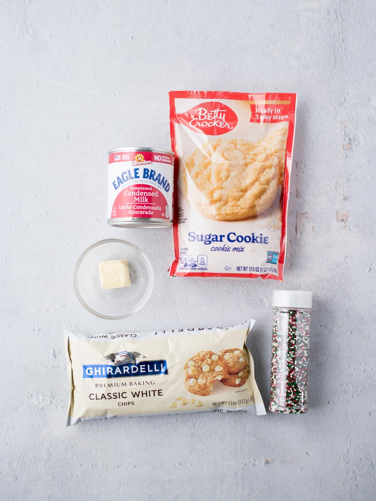 Baking ingredients on a white surface: a bag of sugar cookie mix, a can of condensed milk with a small bowl containing a butter piece, a packet of white chocolate chips, and a container of multicolored sprinkles.