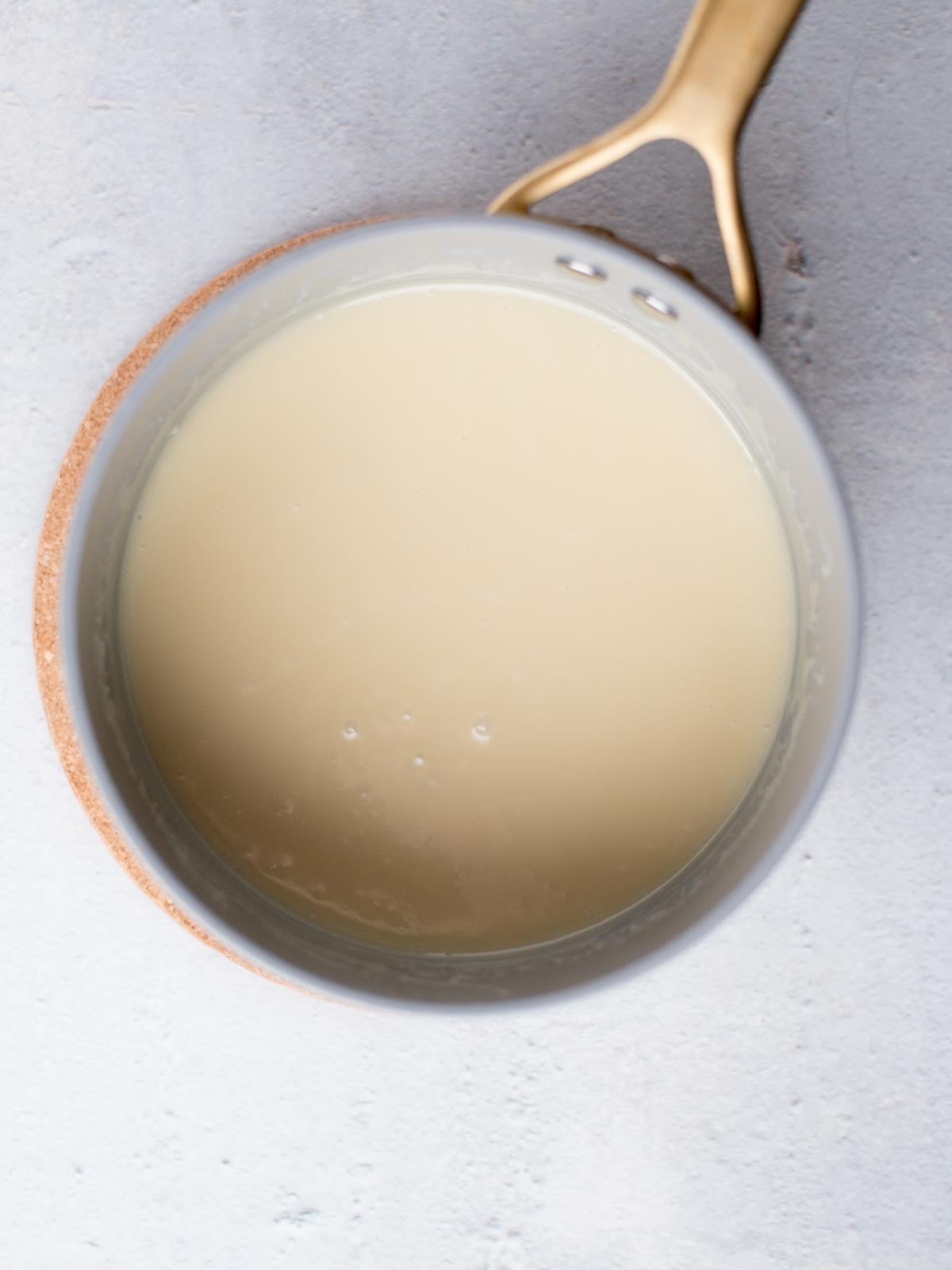 A light-colored sauce in a saucepan with a handle, viewed from above, resting on a light gray surface. The sauces smooth, creamy texture is visible.