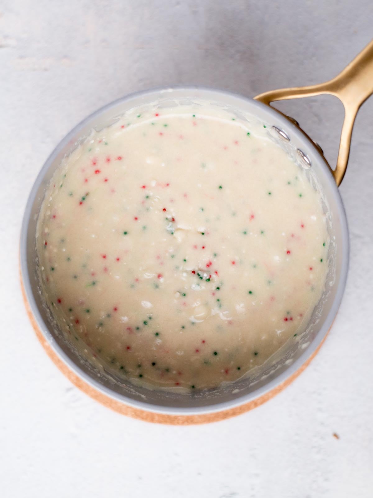 A bowl filled with white cake batter, speckled with small red and green dots, sits on a light surface. A gold-colored handle is visible on the right side.