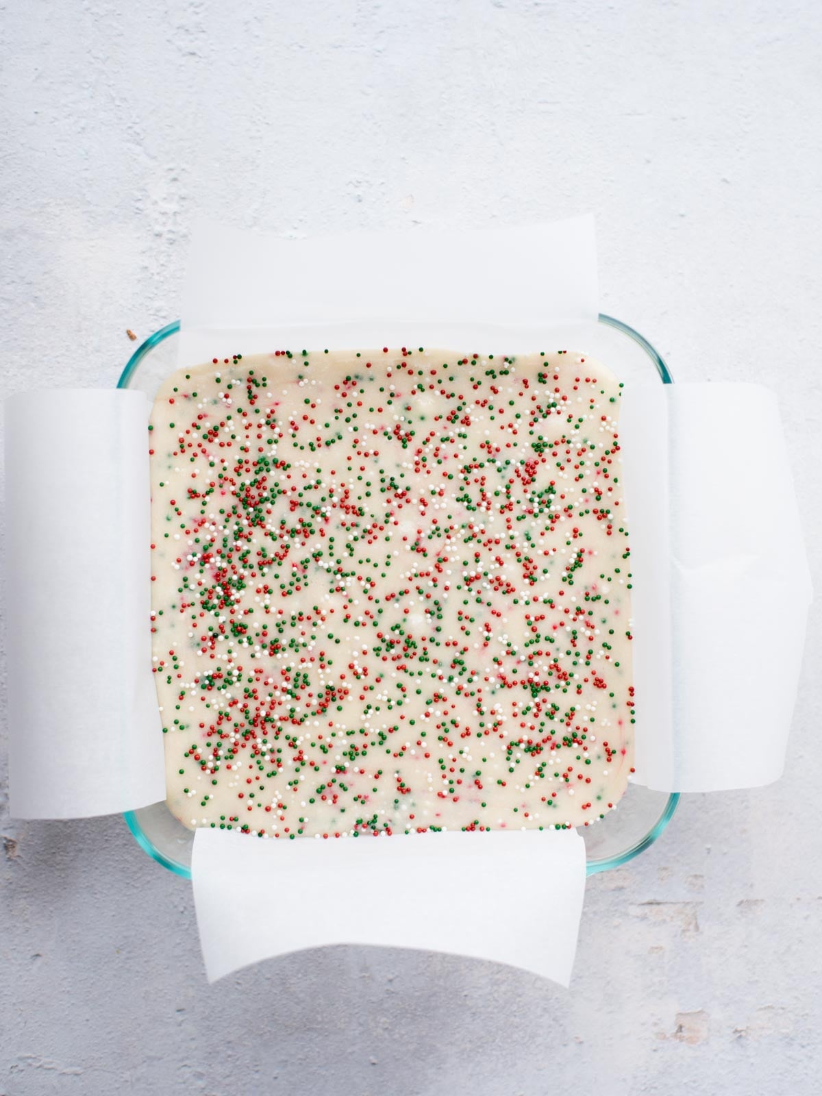 A square glass baking dish lined with white parchment paper is filled with batter topped with red, green, and white sprinkles. The dish is placed on a light-colored surface.