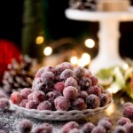 A plate of sugar-coated cranberries sits on a table, surrounded by a festive setting with blurred, glowing lights in the background. Pinecones and foliage add to the holiday ambiance.