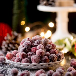 A plate of sugar-coated cranberries sits on a table, surrounded by a festive setting with blurred, glowing lights in the background. Pinecones and foliage add to the holiday ambiance.