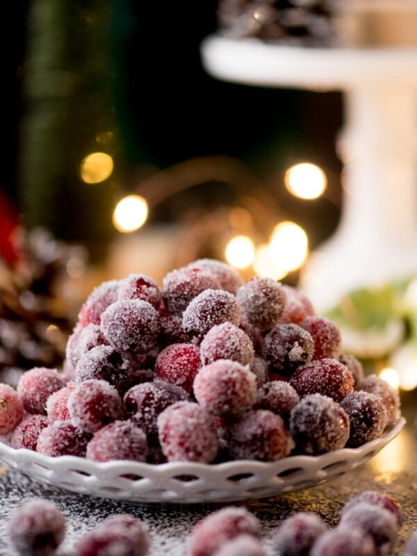 A plate of sugar-coated cranberries sits on a table, surrounded by a festive setting with blurred, glowing lights in the background. Pinecones and foliage add to the holiday ambiance.