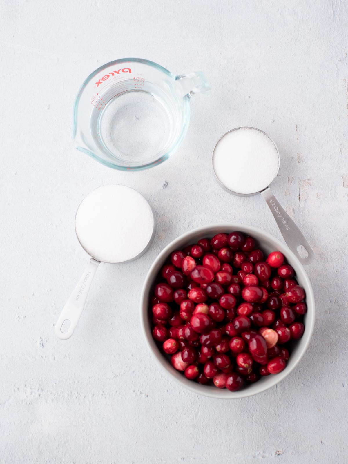 A gray bowl filled with cranberries, a glass measuring cup with water, and two metal measuring cups with sugar are arranged on a light surface.