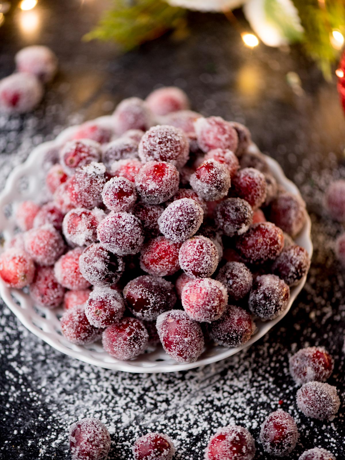 A white bowl is filled with sugared cranberries, surrounded by more cranberries on a dark surface. A dusting of sugar adds a frosty appearance. The background has blurred holiday lights and greenery.