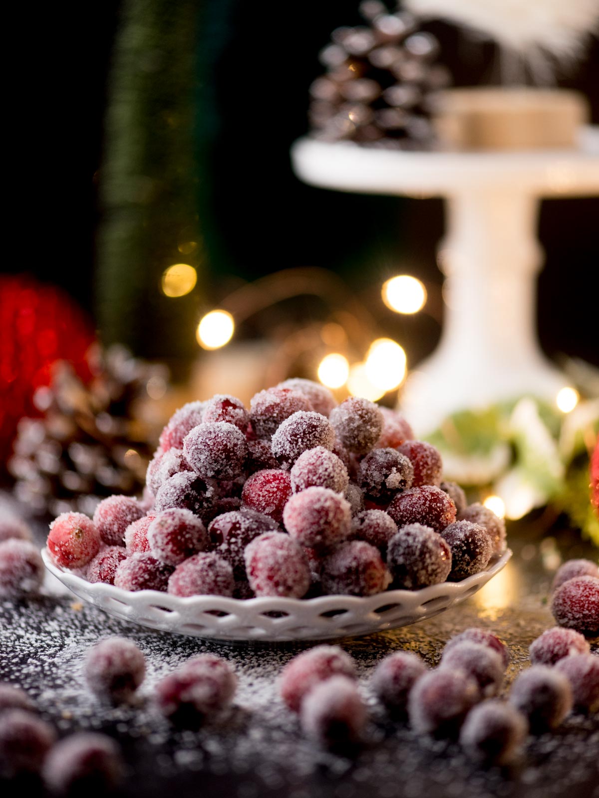 A plate of sugared cranberries sits on a festive table, surrounded by holiday decorations. Pinecones and blurred twinkling lights create a warm, cozy atmosphere in the background.