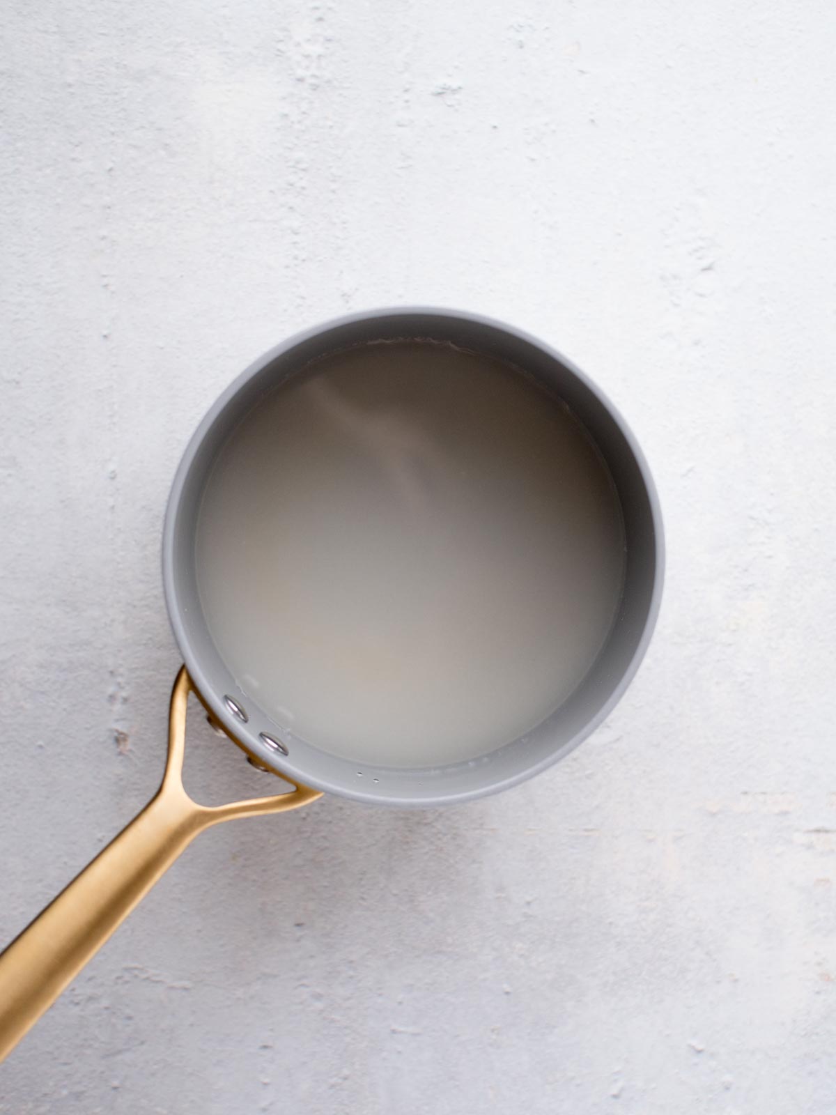 Top-down view of a pot with a gold handle, filled with cloudy water, on a light-colored surface.