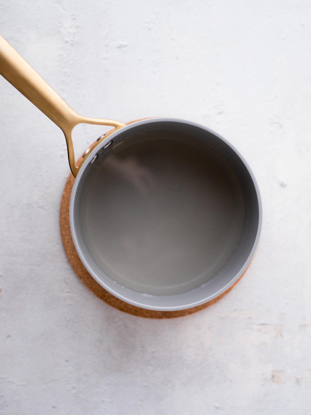A high-angle view of a gray saucepan with a golden handle, placed on a cork trivet. The saucepan is resting on a light textured surface, and it appears to be filled with water.