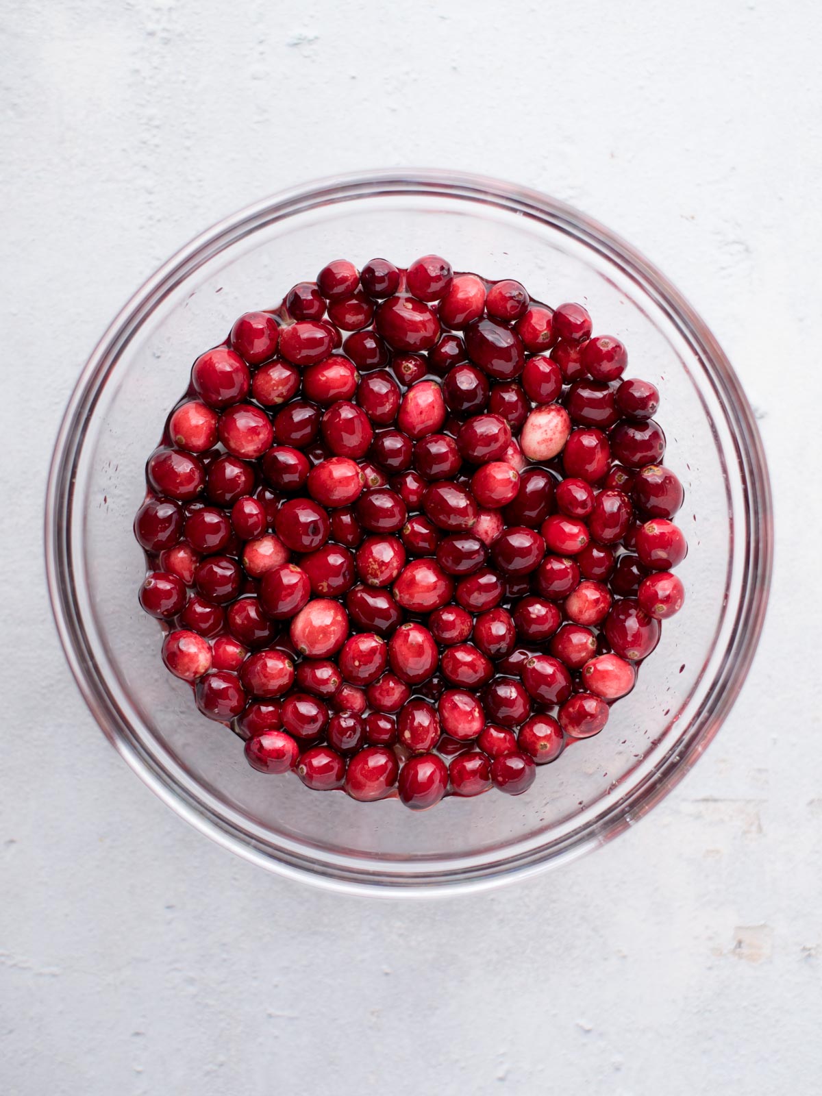 A clear glass bowl filled with vibrant red cranberries, placed on a light gray surface. The cranberries have a glossy appearance, indicating freshness and juiciness.