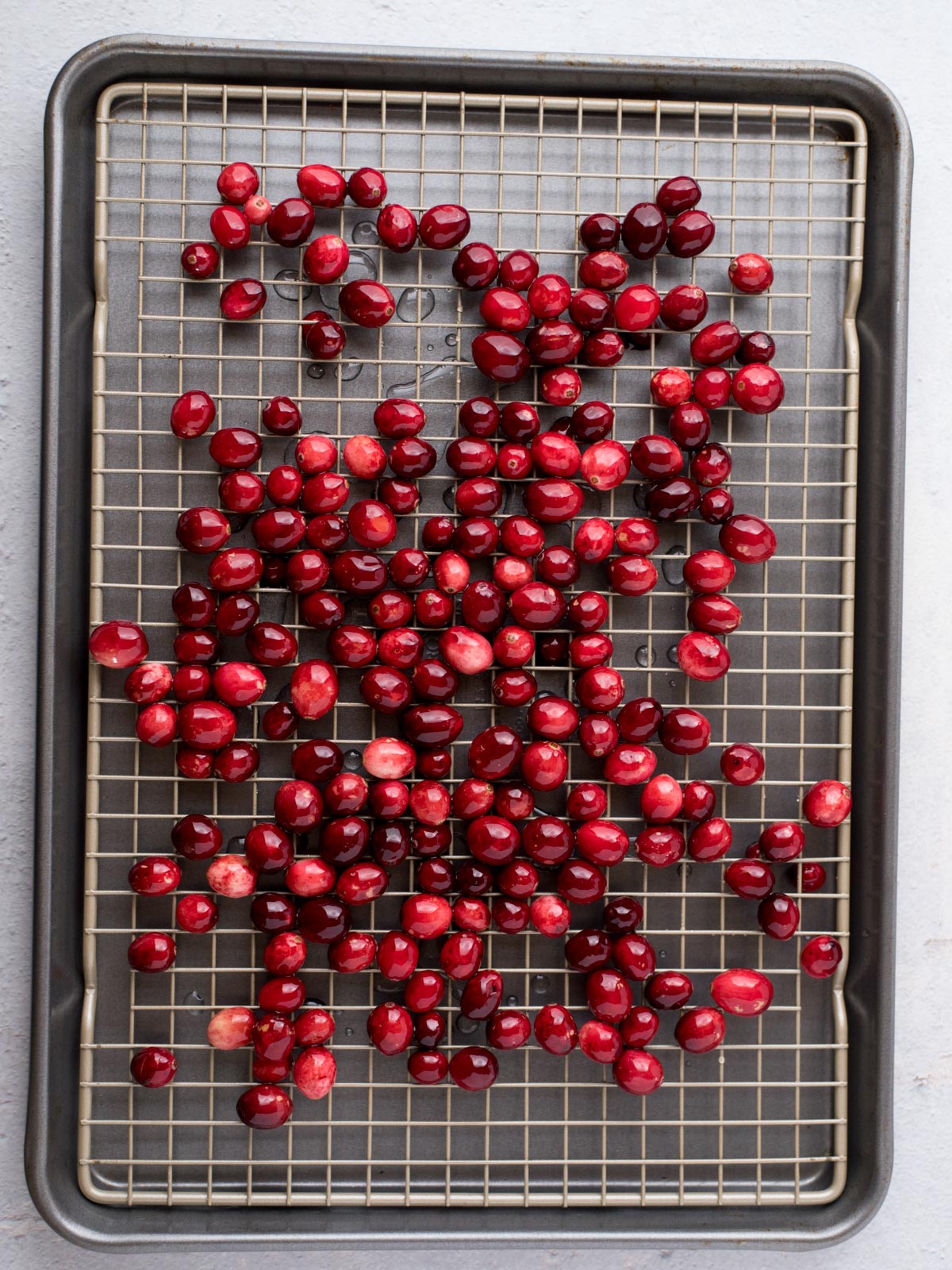 A baking tray with a cooling rack holds a scattered pile of fresh, red cranberries, spread out over the surface. The cranberries vary slightly in size and hue, ranging from bright to deep red. The background is a light, neutral color.