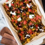 A casserole dish containing a taco bake with ground meat, corn, melted cheese, diced tomatoes, sliced black olives, and dollops of sour cream. The dish is lined with tortillas and garnished with chopped green onions. A wooden serving board is nearby.