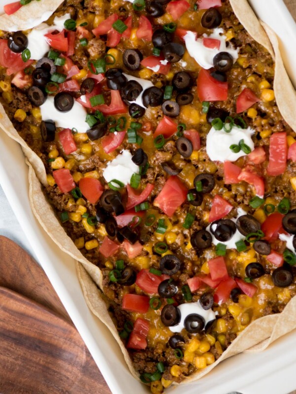 A casserole dish containing a taco bake with ground meat, corn, melted cheese, diced tomatoes, sliced black olives, and dollops of sour cream. The dish is lined with tortillas and garnished with chopped green onions. A wooden serving board is nearby.