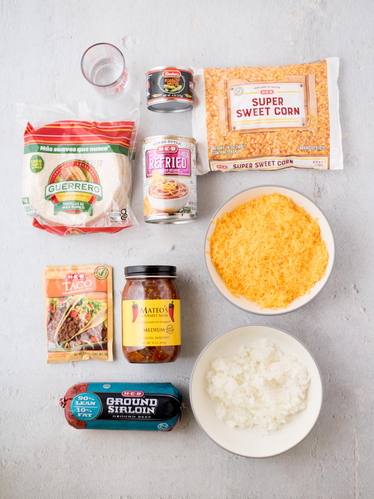 Top view of ingredients on a white surface: corn tortillas, refried beans, taco seasoning mix, salsa, ground sirloin, diced onions, shredded cheese, super sweet corn, and a small glass of water.