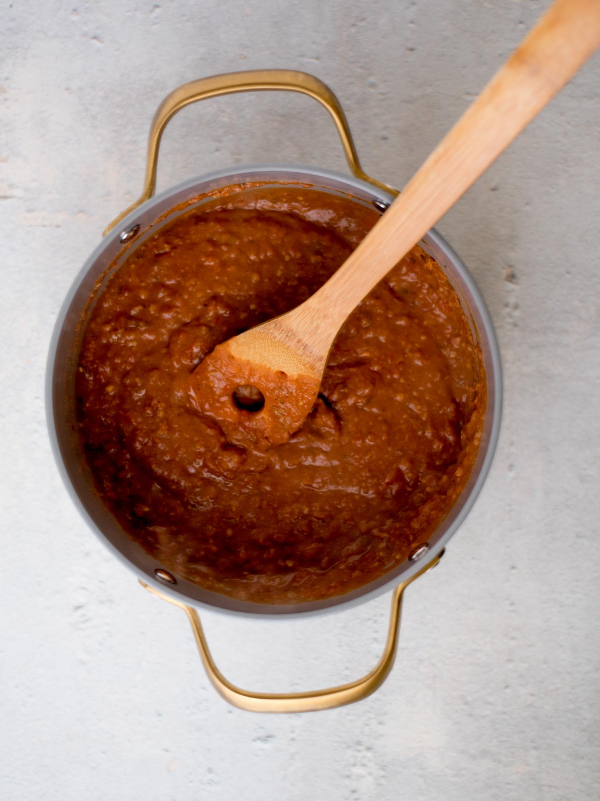 A pot of thick, brown chili with a wooden spoon stirring it, viewed from above. The pot has gold handles and rests on a grey surface. The chili appears rich and textured.