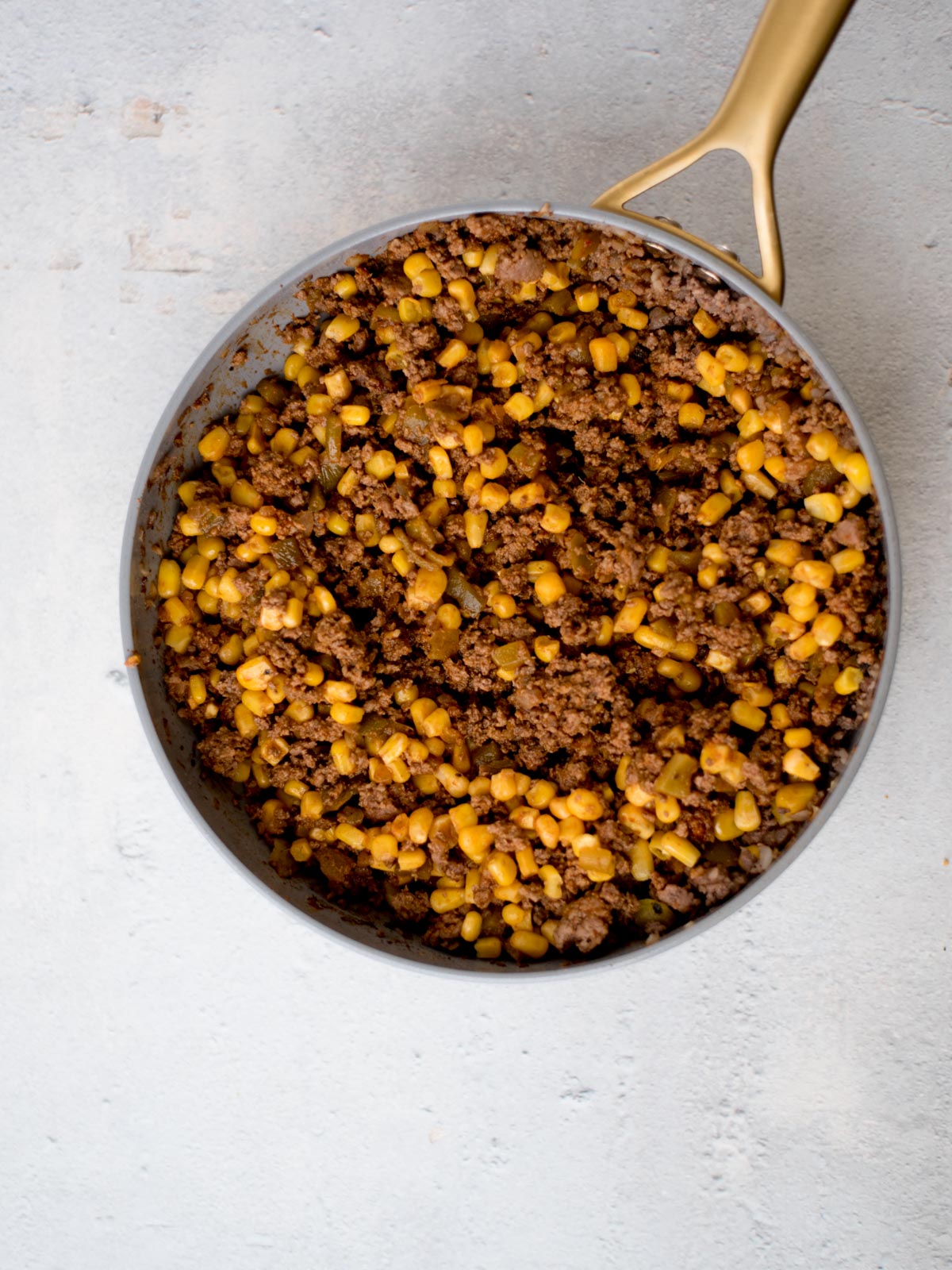 A skillet filled with cooked ground beef and corn kernels on a light gray surface. The ingredients are mixed together, giving a brown and yellow appearance. The skillet has a gold-colored handle.