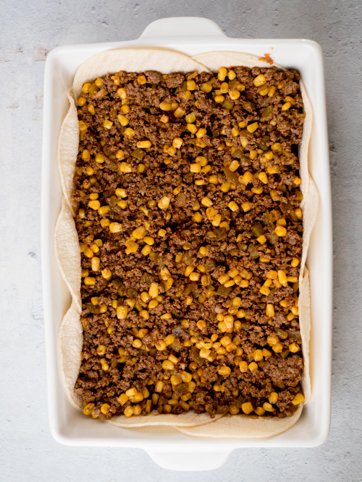 A rectangular baking dish filled with layered tortillas topped with seasoned ground beef and corn. The dish is ready for the oven, resting on a light gray surface.