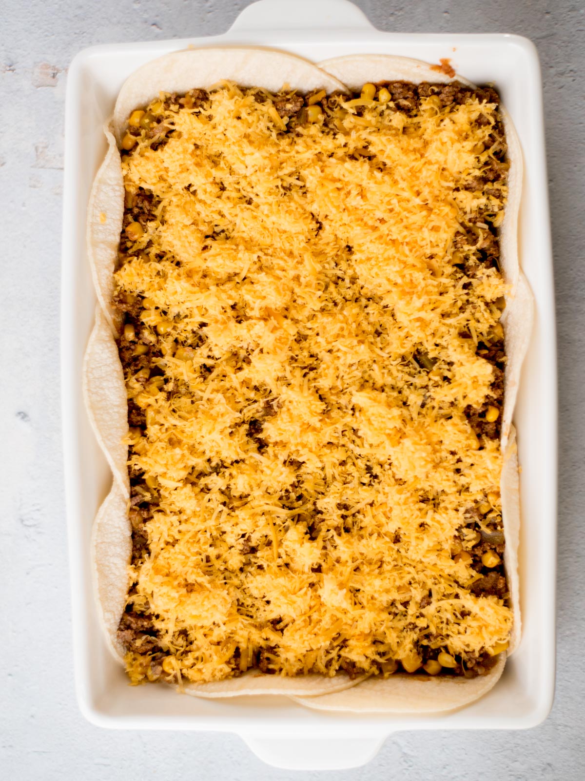 A rectangular baking dish filled with layers of tortillas, ground beef, corn, and melted cheese, creating a casserole. The dish is placed on a light gray surface.