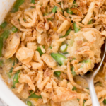 A close-up of a green bean casserole topped with crispy fried onions. A spoonful is being lifted from the white baking dish, revealing creamy sauce and green beans. The dish is on a light-colored cloth with a corner of a napkin visible.