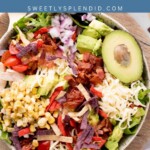 A vibrant Southwest chicken salad graces the bowl, starring grilled chicken, sliced avocado, cherry tomatoes, shredded cheese, corn, crispy bacon, red onion, and crunchy tortilla strips—all elegantly presented on a wooden board.