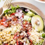 A bowl of Southwest chicken salad brims with ingredients like shredded cheese, corn, tortilla strips, avocado, and diced tomatoes on crisp lettuce. A fork and a plate with chicken sit nearby under the inviting label Southwest Chicken Salad.