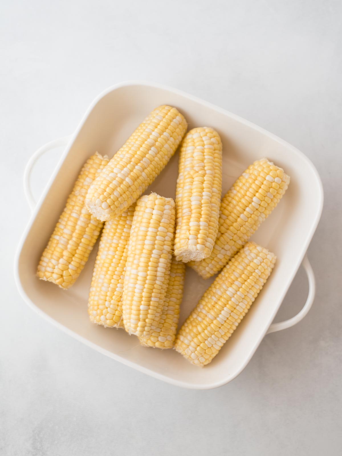A white dish filled with several uncooked, halved ears of corn, neatly arranged on a light-colored surface.